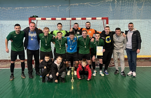 Eine Gruppe von Männern in Medaillen, einer hältend einen Pokal, auf einem Futsal-Platz mit einem Torpfosten und einer Wand im Hintergrund.
