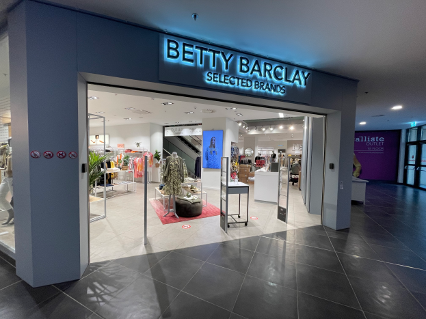 Ein Betty Barclay Selected Brands-Laden in einem Einkaufszentrum mit Schaufensterpuppen in der neuesten Mode, Kämmen mit Kärpern, Tischen mit verschiedenen Gegenständen, Schildern, einer Topfpflanze, Deckenleuchten und einer Tür auf der rechten Seite.