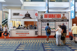 Menschen vor einer Verkaufsbox bei der Nationalen Buchmesse, mit Bannern, Anzeigebildschirm, Treppe, Geländer und Deckenbeleuchtung.