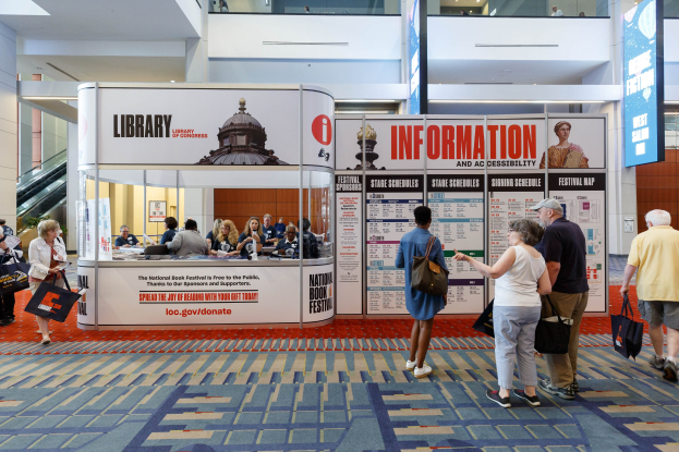 Menschen vor einer Verkaufsbox bei der Nationalen Buchmesse, mit Bannern, Anzeigebildschirm, Treppe, Geländer und Deckenbeleuchtung.