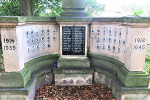 Ein Holocaust-Gedenkmonument in einem jüdischen Friedhof in Berlin, das eine Wand mit Text und Zahlen zeigt, umgeben von Bäumen, einem Zaun und verstreuten trockenen Blättern.