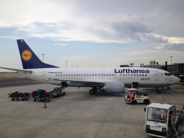 Ein Lufthansa Airbus A320-200 steht am Frankfurter Flughafen, umgeben von verschiedenen Bodenfahrzeugen, Wagen, Verkehrskegeln, Masten und Texttafeln unter einem bewölktem Himmel.