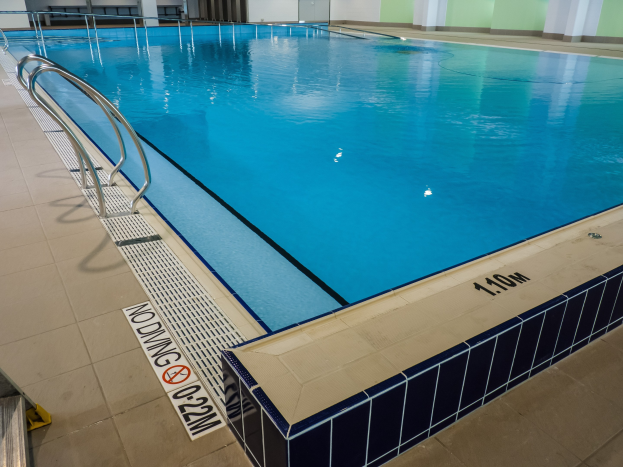 Großes Hallenbad in einem geräumigen Gebäude mit gefliestem Boden, Metallträgern, einer Schautafel, einer Tür, einer Wand und Deckenleuchten.
