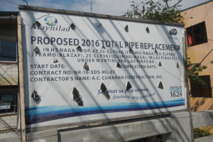 Reklameschild an der Seite eines Gebäudes mit der Aufschrift "Vorgeschlagen 2016 Total Pipe Replacement" im Vordergrund, mit Gebäuden, Bäumen, Drähten und einem klaren blauen Himmel im Hintergrund.