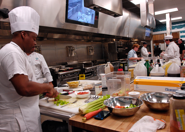 Eine Gruppe von Köchen in einer Küche, die Essen auf einem Schneidbrett zubereiten, mit einem Koch im Vordergrund in einem weißen Kleid und einer Mütze, der ein Messer hält und von verschiedenen Utensilien, Schüsseln, Gemüse, einem Handy, Flaschen und anderen Gegenständen umgeben ist, mit einem Fernseher und Deckenlampen im Hintergrund.