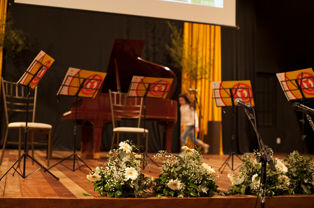 Eine Bühne mit Mikrofonstativen, Blumenbouquets, Stühlen, Notenpulten mit Noten, einem Flügel, einer Person, die läuft, und einem Hintergrund mit Bäumen, Vorhängen und einer Projektionswand.
