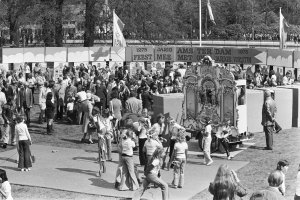 Schwarz-weißes Bild einer Menge, die sich um einen mit Fahnen und Bannern geschmückten Karnevalswagen in einem Park versammelt hat, mit Bäumen und Gebäuden im Hintergrund.