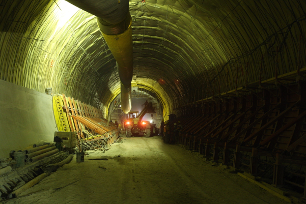 Baustelle mit einem großen Tunnel, Fahrzeugen, verstreuten Holzobjekten, Rohren, einer Wand auf der linken Seite und beleuchteten Lichtern im Hintergrund.
