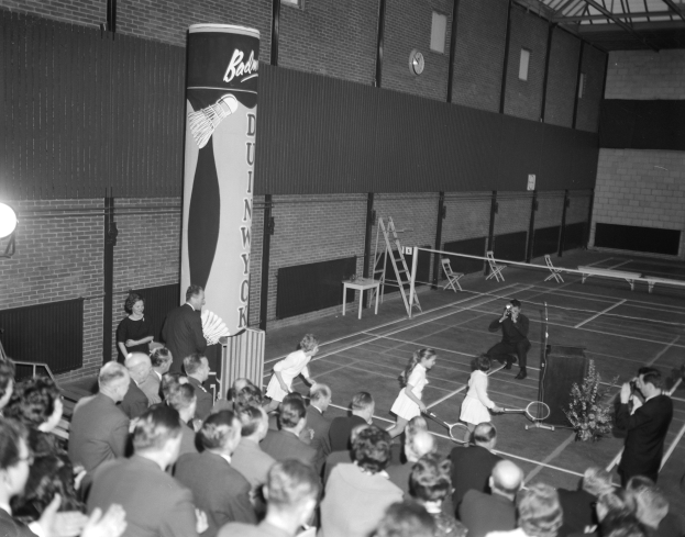 Ein Schwarz-Weiß-Foto einer Gruppe von Menschen, die Tennis in einer Turnhalle spielen, mit einigen Stehenden und anderen auf Stühlen sitzend, die Schläger halten, und einer Uhr an der Wand im Hintergrund.