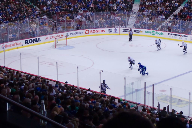 Vier Männer spielen Hockey auf einem Eisstadion, mit zwei stehenden und zwei anderen in Bewegung, einem Netz und einem Zaun dahinter, Zuschauern auf Stühlen im Vorder- und Hintergrund, einem Geländer und einer Treppe in der Ferne.