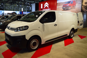 Ein weißer Citroën Berlingo Van wird auf einer Autauschau präsentiert, umgeben von schwarzen Schlafzimmermöbeln, während die Wände mit Brettern und Lichtern verziert sind.