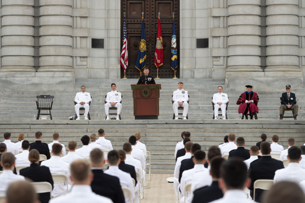 Gruppe von Menschen, die auf Stühlen sitzen und ein Gebäude mit Säulen und einer Tür anschauen, mit einem Podium und Mikrofon in der Mitte, Fahnen im Hintergrund und einem leeren Stuhl links.