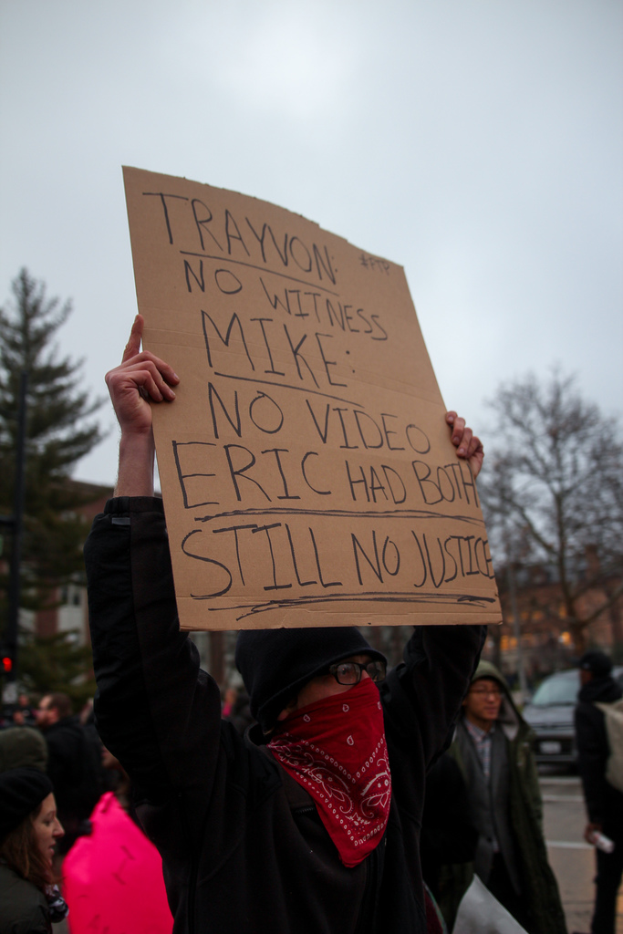 Eine Gruppe von Menschen protestiert auf einer Straße, ein Mann hält ein Schild mit Text, mit Bäumen, einem Auto und einem Himmel im Hintergrund.