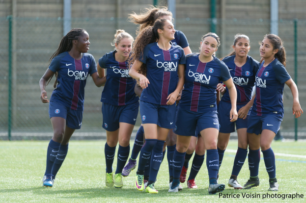 Eine Gruppe junger Frauen, die Fußball auf einem Rasenfeld mit Maschendrahtzaun und einer Wand im Hintergrund spielen, mit der Beschriftung "Paris Saint-Germain Frauenfußball" in der rechten unteren Ecke.