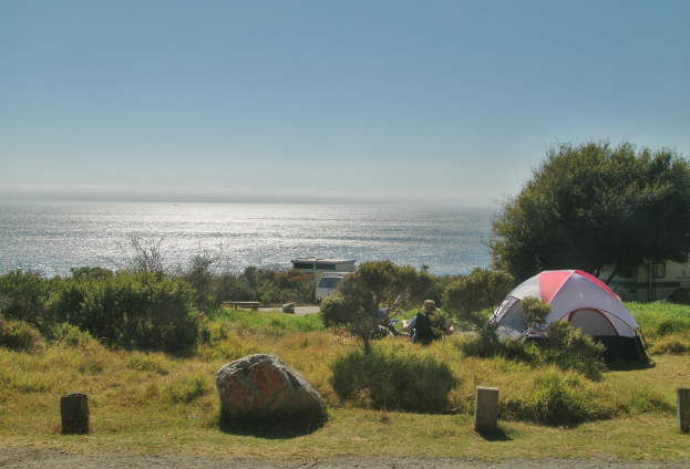 Ein Campingplatz mit einem Zelt, das auf der Seite einer Straße neben dem Ozean aufgestellt ist, mit Menschen, die um das Zelt sitzen, umgeben von Felsen, Pflanzen und Bäumen, mit einem Fahrzeug und der weiten Aussicht auf den Ozean und den Himmel im Hintergrund.