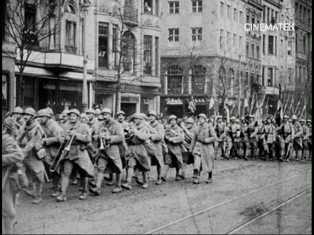 Schwarzes und weißes Foto von Soldaten, die während des Ersten Weltkriegs eine Straße in Berlin entlangmarschieren, mit Mützen und Gewehren, sowie Bäumen und Gebäuden an der Straße und einem Pfahl im Hintergrund.