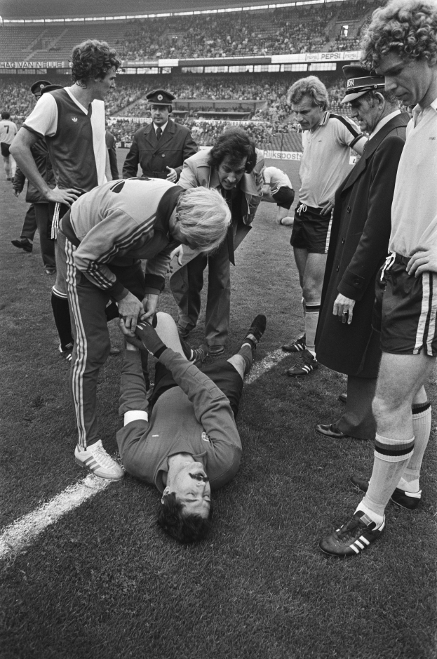 Schwarzes und weißes Bild eines Füßballspielers, der auf dem Boden liegt und von einer Gruppe von Männern umgeben ist, mit einer Menschenmenge im Hintergrund eines Stadions.