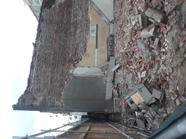 Teilweise abgerissenes Gebäude mit verstreuten Ziegeln und Metallstangen, mit anderen Gebäuden und einem klaren blauen Himmel im Hintergrund.