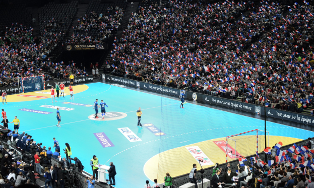 Eine Gruppe von Menschen, die Handball spielen, in einem Stadion mit einem Netz, das das Spielfeld teilt, und Zuschauern in den Rängen, während Fotografen das Spiel aufnehmen.