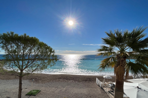 Ein Strand mit Palmen, Sonnenschirmen, üppiger Vegetation und einem strahlend blauen Himmel mit der Sonne im Hintergrund.