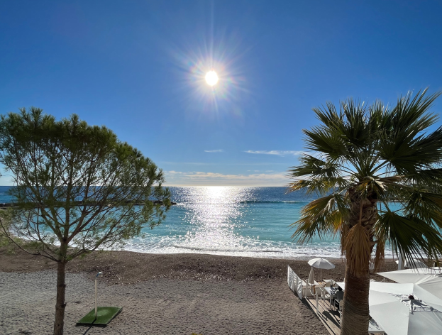 Ein Strand mit Palmen, Sonnenschirmen, üppiger Vegetation und einem strahlend blauen Himmel mit der Sonne im Hintergrund.