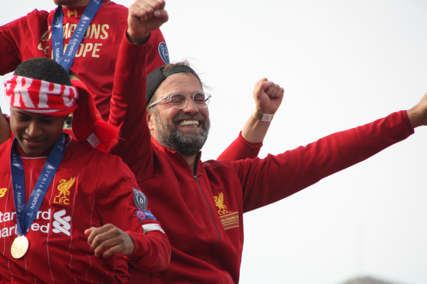 Liverpool-Trainer Jürgen Klopp feiert mit seinen Spielern den Sieg in der UEFA Champions League, während zwei medaillentragende Personen in der Nähe sind und der Himmel im Hintergrund zu sehen ist.