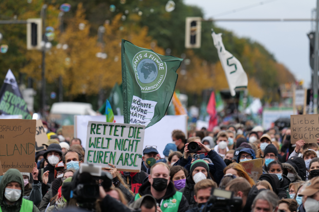 Eine große Gruppe von Menschen mit Masken und Schildern, Fahnen und Kameras nimmt an einem Klimastreik in Deutschland teil, umgeben von Bäumen, Pfosten, Verkehrszeichen und Fahrzeugen, mit dem Himmel im Hintergrund.