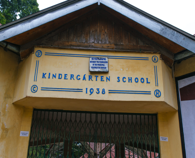 Gelbes Gebäude mit einer "Kindergarten-Schule"-Schilder und einer Namenstafel an der Wand, ein Gitter unten, Bäume im Hintergrund und einen klaren blauen Himmel.