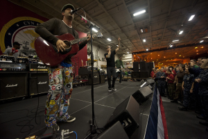 Ein als Chris Stapleton identifizierter Mann spielt eine akustische Gitarre auf der Bühne, trägt eine Mütze, mit einem Mikrofon, Lautsprechern, einer Fahne und anderen Instrumenten im Hintergrund, vor einem Publikum unter Deckenlampen mit einem Banner an der Wand dahinter.