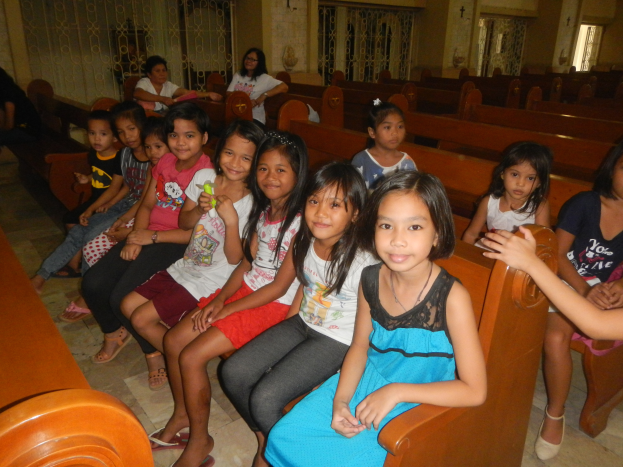 Eine Gruppe junger Mädchen sitzt auf Kirchenbänken, mit einigen Menschen im Hintergrund, umgeben von Wänden mit Gittern und Fenstern.