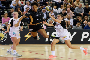 Eine Gruppe von Frauen, die Handball auf einem Platz spielen, mit einem Ball in der Mitte, während Zuschauer im Hintergrund sitzen und ein Banner mit Text und Logos zu sehen ist.