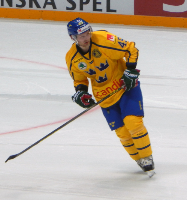 Ein Eishockeyspieler in Bewegung auf dem Eis, der einen Helm, Handschuhe und Schlittschuhe trägt und einen Hockeystock hält, mit einer Tafel im Hintergrund mit Text.