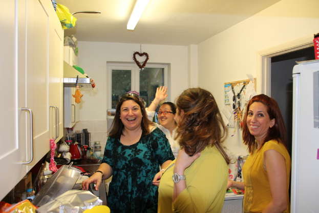 Eine Gruppe strahlender Frauen in einer Küche, eine trägt eine Brille, mit einem Kühlschrank, Gefäßen auf der Arbeitsplatte, Schränken und einer Wandtafel an der Wand, unter einer Deckenlampe.