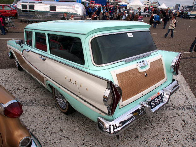 Ein grüner und weiße Kombiwagen auf einem belebten Parkplatz mit Zelten, Masten, Bäumen und einem klaren blauen Himmel im Hintergrund.