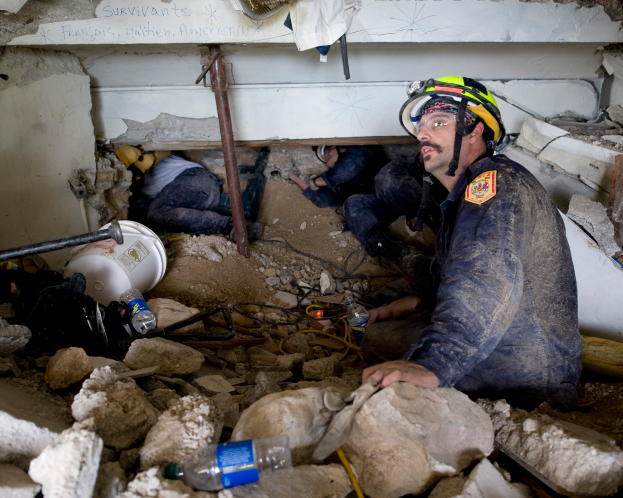 Ein Feuerwehrmann mit Helm sitzt zwischen Trümmern, darunter Steine und Flaschen, mit einer teilweise sichtbaren Wand im Hintergrund, die wahrscheinlich den Namen des Gebäudes trägt, das Schauplatz eines israelischen Luftangriffs ist.