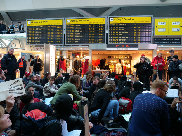 Eine große Gruppe von Menschen sitzt und steht in einem Flughafen während einer Protestaktion, mit Gepäck und Papieren in Sicht, während Anzeigetafeln, Schaufensterpuppen in Kleidern und Deckenleuchten im Hintergrund zu sehen sind.