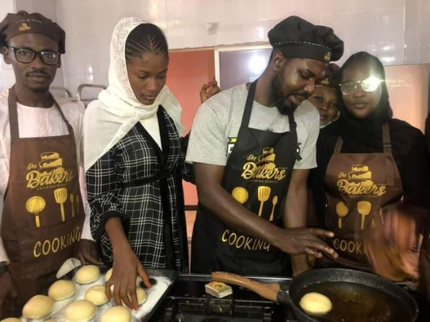 Eine Gruppe von Menschen in Schürzen und Mützen, die in einer Küche mit einem Topf auf dem Herd, Schränken und einer Wand im Hintergrund Essen zubereiten.