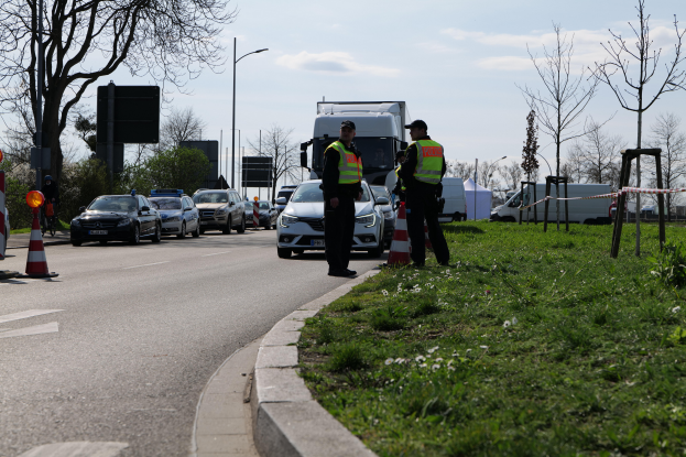 Zwei Polizisten stehen an der Seite einer Straße neben einem Auto, mit Verkehrskegeln, Gras, Pflanzen, Bäumen, Pfosten, Schildern und anderen Gegenständen im Vordergrund und einem bewölkten Himmel im Hintergrund.