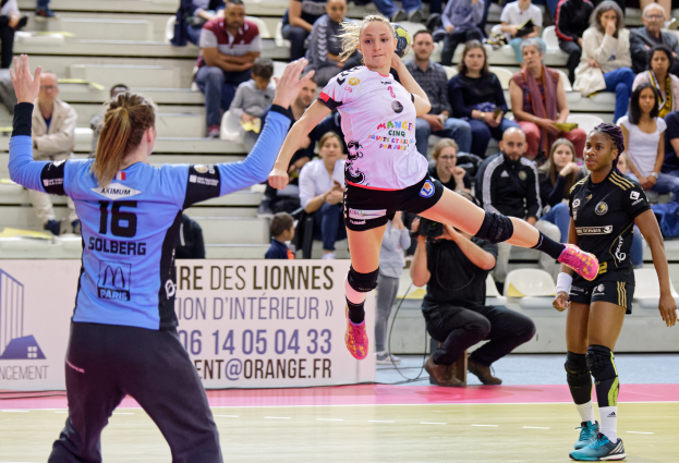 Eine Gruppe Frauen beim Handballspielen auf einem Court mit verstreuten Schuhen, während eine Menge von Stühlen und Treppen aus zusieht und eine Person das Ereignis mit einer Kamera filmt.