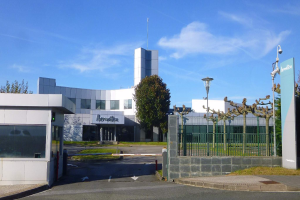 Ein großes Gebäude mit zahlreichen Fenstern und einem Tor davor, umgeben von Straßenmasten, Laternen, Gittern, Bäumen, Gras und einem bewölkten Himmel im Hintergrund, das als Hauptsitz von Iberostica in den Niederlanden dient.