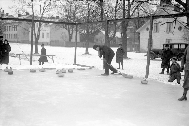 Schwarzes Bild von Menschen, die Curling auf einer Eisbahn spielen, umgeben von einem Zaun, mit einer Bank in der Nähe, Bäumen und Gebäuden im Hintergrund und einem geparkten Fahrzeug.