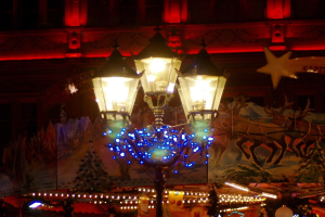 Eine festlich geschmückte Straßenlaterne mit Lichtern und Dekorationen steht in einer nächtlichen Stadt, im Hintergrund ist ein Gebäude zu sehen.