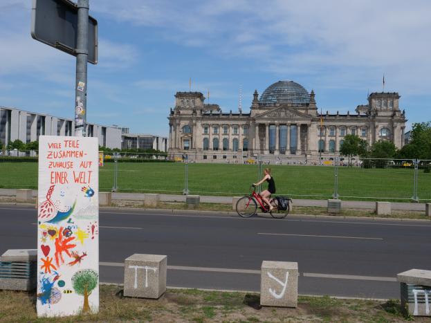 Eine Person in einem schwarzen Kleid fährt mit dem Fahrrad die Straße vor dem Reichstag in Berlin, Deutschland, entlang, mit Gebäuden, Bäumen und Menschen im Hintergrund unter einem blauen und weißen Himmel.
