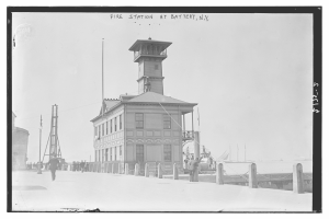 Schwarzes und weißes Foto einer Feuerwache an der Battery, New York, mit einem Gebäude, Menschen, Pfählen, einem Zaun, Booten auf dem Wasser und einem bewölkten Himmel.