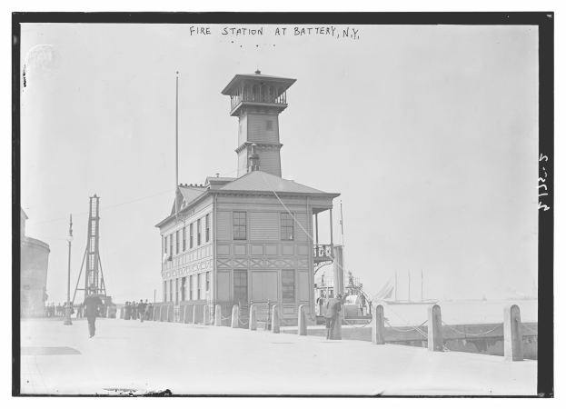 Schwarzes und weißes Foto einer Feuerwache an der Battery, New York, mit einem Gebäude, Menschen, Pfählen, einem Zaun, Booten auf dem Wasser und einem bewölkten Himmel.