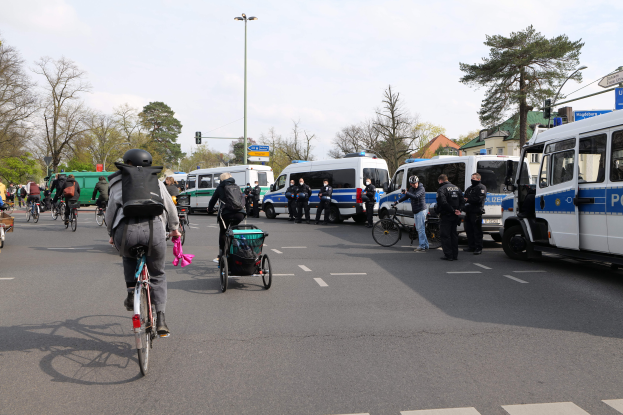Eine Gruppe von Menschen, die auf Fahrrädern eine von Polizeiwagen und anderen Fahrzeugen gesäumte Straße entlangfahren, einige tragen Helme und Taschen, andere schieben Kinderwagen, mit Bäumen, Laternenmasten, Verkehrsampeln, Schildern, Gebäuden und einem bewölkten Himmel im Hintergrund.