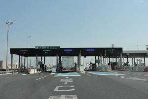 Mautstation an einer Straßenrand mit einem geparkten Lkw, weißen und blauen Straßenmarkierungen, Laternenmasten, Schildern und fernen Gebäuden, Bäumen und einem klaren Himmel.