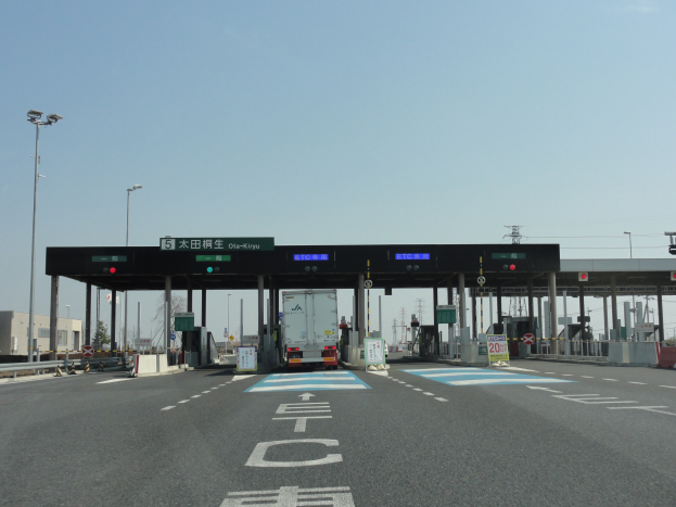 Mautstation an einer Straßenrand mit einem geparkten Lkw, weißen und blauen Straßenmarkierungen, Laternenmasten, Schildern und fernen Gebäuden, Bäumen und einem klaren Himmel.