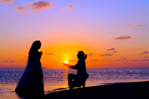 Bräutigam und Braut stehen am Strand bei Sonnenuntergang, wobei der Bräutigam in der Hocke sitzt und ein Objekt hält, die Braut in einem weißen Kleid, vor einem Himmel aus Orangen, Pinks und Purpur und tiefem blauem Wasser.