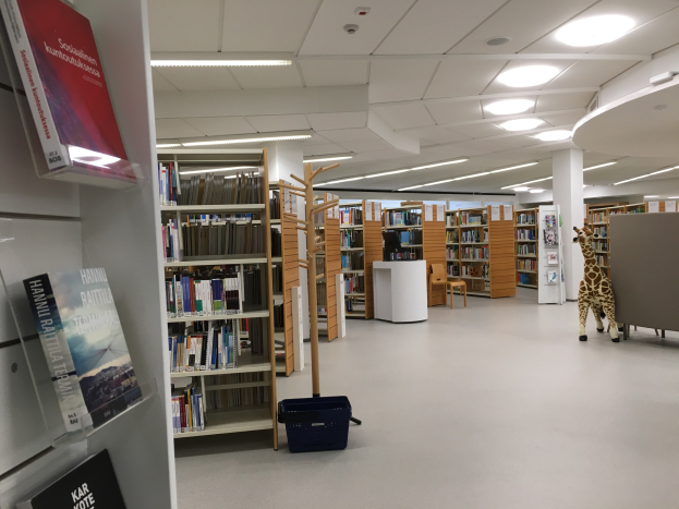 Ein geräumiger Bibliotheksraum mit hohen Bücherregalen, gefüllt mit Büchern, einem Korb auf dem Boden, einem Giraffen-Stofftier, Stühlen, einem Tisch, tragenden Säulen und Deckenleuchten.